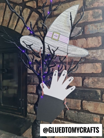 A Halloween craft features a black tree branch with a paper witch hat hot air balloon and a white hand-shaped cutout hanging from it. The background shows a brick wall. @GLUEDTOMYCRAFTS is written at the bottom.