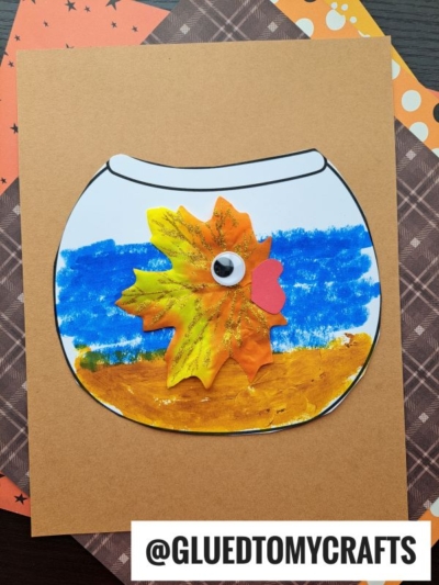 A paper fishbowl craft featuring a leaf fish with glitter, a googly eye, blue and yellow paint accents, all displayed on brown paper with patterned backgrounds.