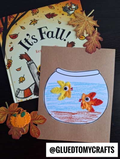 A children’s fall craft with a paper fishbowl drawing featuring leaf fish made from autumn leaves, placed on a table with a fall-themed book, faux leaves, and a pumpkin cutout. Text reads @GLUEDTOMYCRAFTS.
