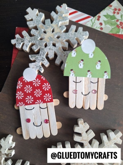 Two Christmas crafts, crafted as cheerful craft stick marshmallow friends with red and green holiday hats and pink cheeks, are displayed on a dark surface alongside wooden snowflakes and festive paper.