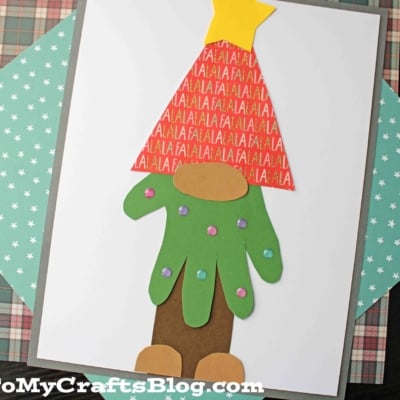 A Christmas craft featuring a handprint Christmas tree gnome made from cut-out paper shapes: a Fa La La patterned hat with a yellow star, a green glove-like beard with gems, and brown boots, arranged on a white paper background.