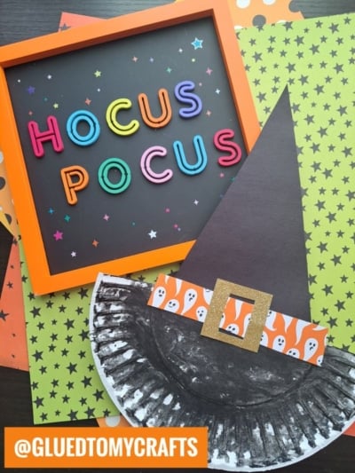 Colorful Hocus Pocus sign in an orange frame sits above a charming paper plate witch hat craft, featuring a ghost-patterned band, all displayed on festive star and polka dot paper, plus @GLUEDTOMYCRAFTS label in orange.