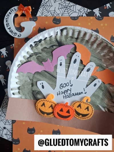 A Halloween craft features handprint ghosts, a pink bat, felt pumpkins, and a painted paper plate designed as a charming paper plate pumpkin patch. The tag @GLUEDTOMYCRAFTS is included amid festive backgrounds with cats and bats, making this pumpkin patch craft perfect for the spooky season.