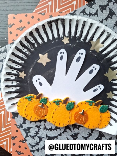 A Halloween craft featuring a black-painted paper plate with a white paper handprint ghost, gold star stickers, and cut-out pumpkins with green leaves—perfect for creating your own haunted pumpkin patch with patterned orange and black backgrounds.
