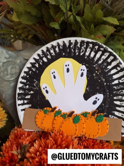 A paper plate pumpkin patch craft features a black painted rim, a yellow circle, and a white paper hand with ghost faces. Three felt pumpkins and orange flowers sit at the bottom, with the text @GLUEDTOMYCRAFTS.