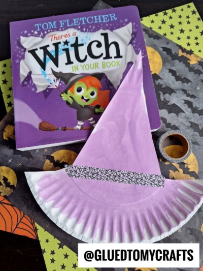 A children’s book titled There’s a Witch in Your Book sits on a Halloween-themed surface next to a homemade paper plate witch hat, crafted from a purple plate and decorated with washi tape. A roll of black-patterned tape is nearby, completing the spooky scene.