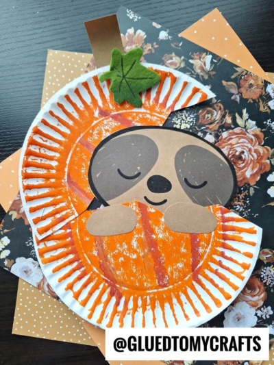 A cute paper plate sloth in pumpkin craft made from a paper plate, orange paint, and colored paper, lying on floral and polka dot paper backgrounds. The sloth appears to be sleeping, cuddling a pumpkin. The handle @GLUEDTOMYCRAFTS is shown.