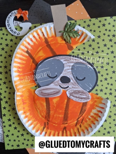 A paper plate craft features a smiling paper plate sloth in pumpkin, with a gray face, brown nose, and closed eyes, nestled in orange paint and surrounded by colorful paper sheets. Text reads @GLUEDTOMYCRAFTS.