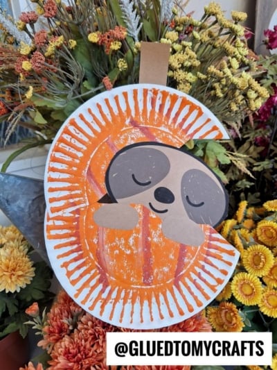 A cute paper plate sloth in pumpkin craft, made from a painted plate and paper cutouts, hangs among colorful autumn flowers. The sloth looks cozy and sleepy, with its head and arms attached. Text: @GLUEDTOMYCRAFTS.