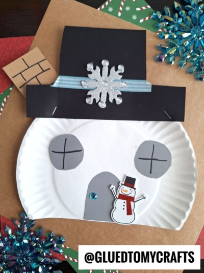 A paper plate snowman house craft, featuring a black paper hat with a glittery snowflake, paper chimney, ribbon, and sticker snowman, is displayed on a festive background adorned with blue snowflakes.