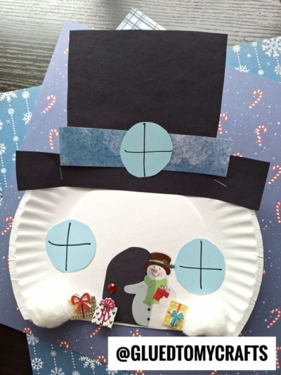 A paper plate snowman face features blue paper eyes, a large black hat, cotton ball cheeks, and small Christmas presents with a snowman sticker at the bottom—perfect decor for your paper plate snowman house against candy cane patterned paper.