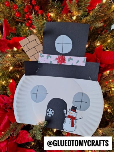 A paper plate snowman house face, crafted with black paper shapes, features a drawn snowman, snowflake, and chimney, hanging on a Christmas tree with red ornaments. The handle @GLUEDTOMYCRAFTS is shown.