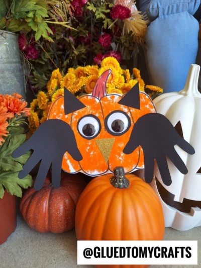 A pumpkin-shaped craft with googly eyes, handprint wings, and a yellow beak sits among real pumpkins and flowers, capturing the whimsical charm of a fall owl craft. The username @GLUEDTOMYCRAFTS is displayed at the bottom.