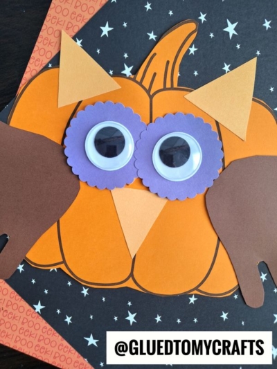 A Halloween craft featuring a paper pumpkin owl decorated with large googly eyes, purple scalloped eye frames, tan and brown paper ears, and a black star-patterned background. @GLUEDTOMYCRAFTS text is at the bottom.