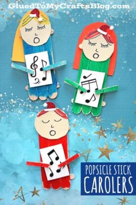 Three colorful popsicle stick Christmas carolers with paper faces and music sheets are arranged on a blue starry background. Each figure has a small Santa hat and a mini clothespin arm holding a song sheet.