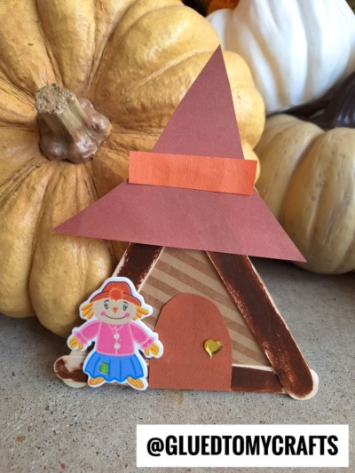 A craft stick scarecrow house with a paper hat, a yellow heart on the door, and a sticker of a girl stands in front of pumpkins. Text reads @GLUEDTOMYCRAFTS.