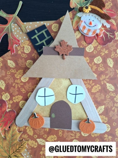A triangular popsicle stick scarecrow house decorated with pumpkins, windows, and a leaf; fall leaves and a smiling scarecrow background; @GLUEDTOMYCRAFTS label at the bottom.
