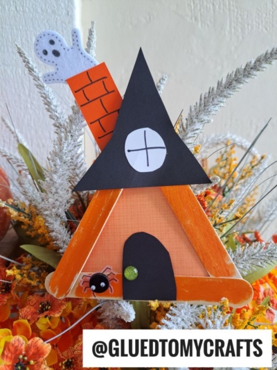 A popsicle stick witch house craft for Halloween, featuring orange sticks shaped into a triangle with a black roof, a spooky paper ghost in the chimney, and floral decorations. Text reads @GLUEDTOMYCRAFTS.