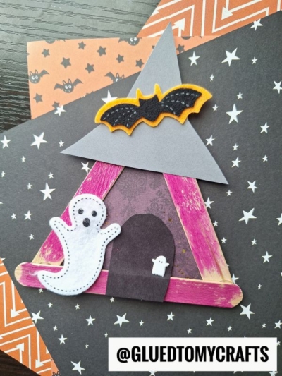 A Halloween craft featuring a popsicle stick witch house, with ghost and bat decorations, placed on black and orange star-patterned papers. The Instagram handle @GLUEDTOMYCRAFTS is at the bottom.