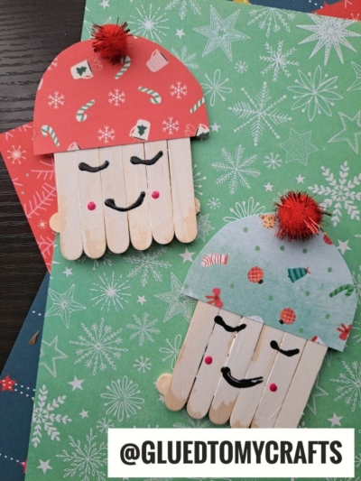 Two festive craft stick marshmallow friends, made from popsicle sticks, colored paper, and pom-poms, look like smiling faces in winter hats. They sit on green and red holiday-themed paper. Text reads @GLUEDTOMYCRAFTS.