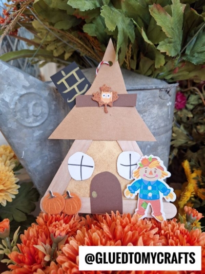 A handmade scarecrow house craft shaped like a teepee, decorated with pumpkins, a small scarecrow figure, fall leaves, and orange flowers. A tag reads @GLUEDTOMYCRAFTS.