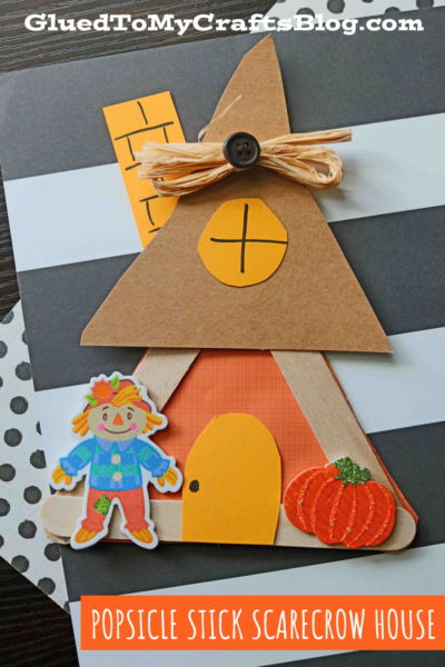 A popsicle stick scarecrow house decorated with a paper door, scarecrow sticker, pumpkin, and a brown triangle roof topped with raffia and a button. Text reads POPSICLE STICK SCARECROW HOUSE. Fun and easy scarecrow house craft!.
