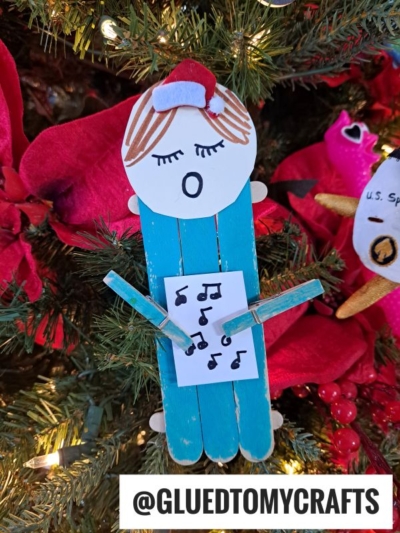 A handmade popsicle stick Christmas carolers ornament shaped like a caroler, made from blue popsicle sticks, paper, and clothespins, hangs on the tree holding music notes and wearing a red Santa hat.