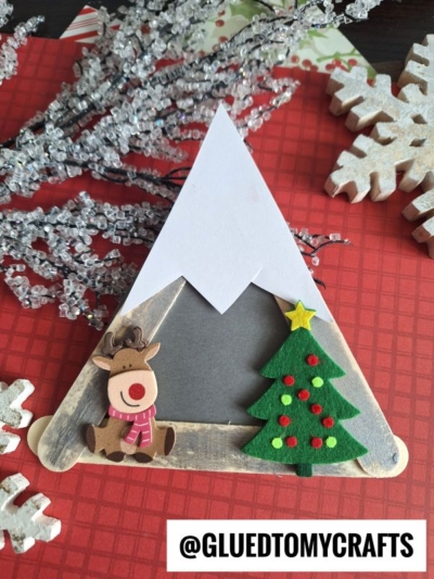 A handmade holiday craft shaped like a popsicle stick snowy mountain, decorated with a felt Christmas tree, reindeer, and snow, displayed on a red surface with snowflake ornaments and silver branches nearby.