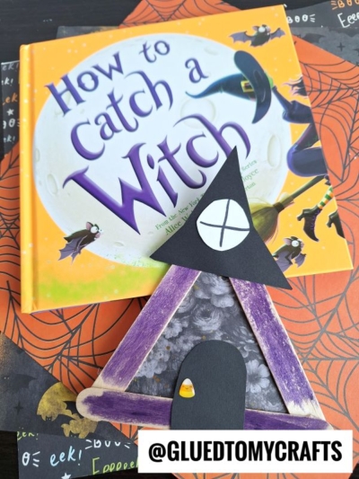 A Halloween children’s book titled “How to Catch a Witch” sits on decorative paper, with a popsicle stick witch house craft and a purple witch hat made from painted sticks and paper placed in front. Text at the bottom reads @GLUEDTOMYCRAFTS.