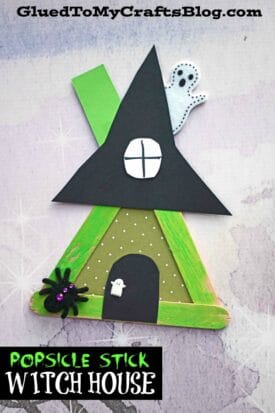 A popsicle stick witch house craft featuring a black hat, green and black accents, a ghost, spider, door, and white polka dots, all displayed on a light background. Text reads: Popsicle Stick Witch House.