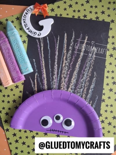 Vibrant Chalk Art Monster crafted from a purple paper plate, featuring googly eyes and pastel chalk crayons. The monster sits atop black paper adorned with colorful chalk fireworks on a star-patterned background. @GLUEDTOMYCRAFTS is written at the bottom.