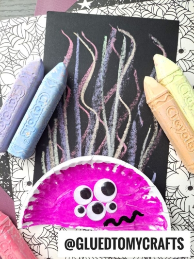 A colorful Chalk Art Monster jellyfish craft made from a painted paper plate with googly eyes, set on black paper with pastel chalk lines as tentacles, surrounded by large Crayola chalk sticks. @GLUEDTOMYCRAFTS label at the bottom.