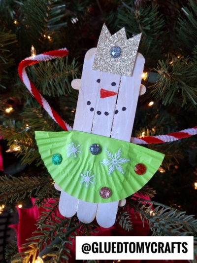 A handmade popsicle stick snowman ballerina ornament features a silver glitter crown, green cupcake liner skirt with sequins, and pipe cleaner arms, hanging gracefully on a Christmas tree—a delightful snowman ballerina craft.