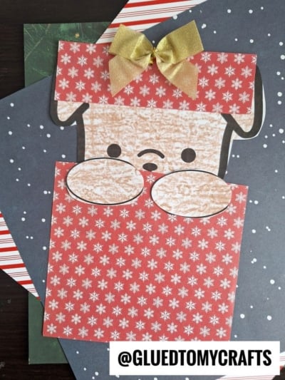 A handmade Peeking Christmas Puppy card with a red and white snowflake pattern, a gold bow on its head, and a sad face. The background shows layered festive paper and the handle @GLUEDTOMYCRAFTS at the bottom.