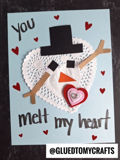 This doily snowman card features a snowman crafted from a white paper heart doily, felt hearts, and paper cutouts for details. Red hearts and “You melt my heart” decorate the blue background for a sweet handmade touch.
