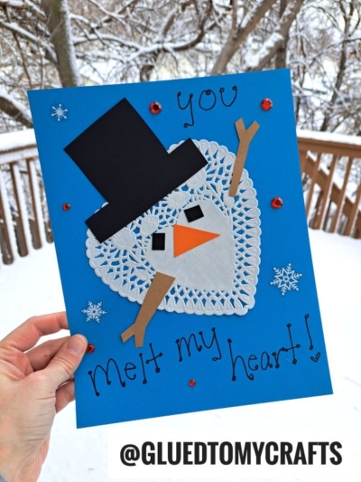 A hand holds a blue doily snowman card with a snowman made from a white heart paper doily, black hat, orange nose, and brown stick arms. Text reads "You melt my heart!" Snow and trees are visible outside. @GLUEDTOMYCRAFTS is printed below.