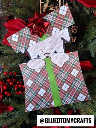 A Peeking Christmas Puppy craft hangs on the tree—made from plaid-patterned paper, shaped like a present with a green ribbon. The dog has googly eyes, a sad mouth, and a red bow. @GLUEDTOMYCRAFTS is featured at the bottom.
