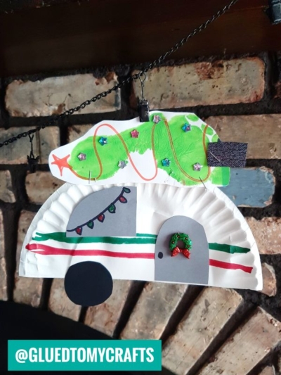A handmade paper plate Christmas camper decoration made from colored paper and markers features a green Christmas tree on top with sequin ornaments, festive lights, and a wreath, all hanging in front of a brick wall.