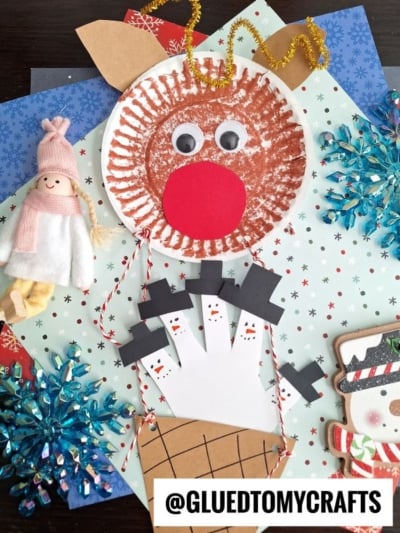 A festive craft display featuring a paper plate reindeer hot air balloon, a handprint snowman, blue snowflakes, a small doll in a pink hat, and a snowman drawing—all arranged on patterned paper. @GLUEDTOMYCRAFTS label at the bottom.