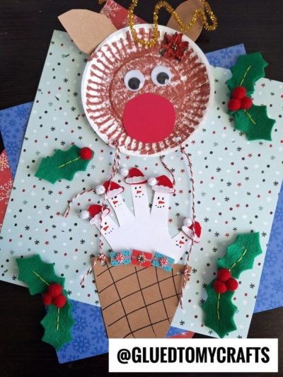 A festive reindeer hot air balloon craft made from a paper plate, brown coloring, a red paper nose, handprint cutouts, and holiday decorations—all arranged on themed paper with felt holly accents. @GLUEDTOMYCRAFTS.
