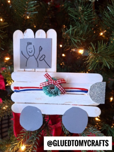 A Christmas tree ornament made from popsicle sticks resembles a white popsicle stick mail truck with a gray window showing a stick-figure drawing. The truck is decorated with ribbon, a mini wreath, and gray wheels, while Christmas lights shine in the background.