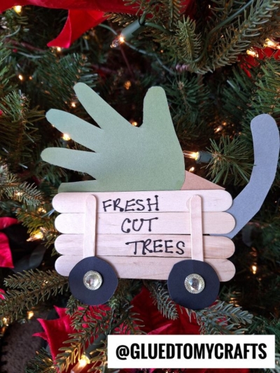 A handmade popsicle stick Christmas tree wagon, crafted with black and white paper and green and gray hand-shaped cutouts, labeled FRESH CUT TREES, is displayed in front of a decorated Christmas tree.