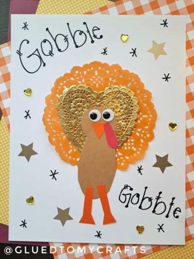 A handmade paper doily turkey craft features orange and gold doilies, googly eyes, and paper cut-outs on white paper. “Gobble Gobble” is written in black, surrounded by gold stars and hearts—perfect for creating festive paper doily turkey cards.