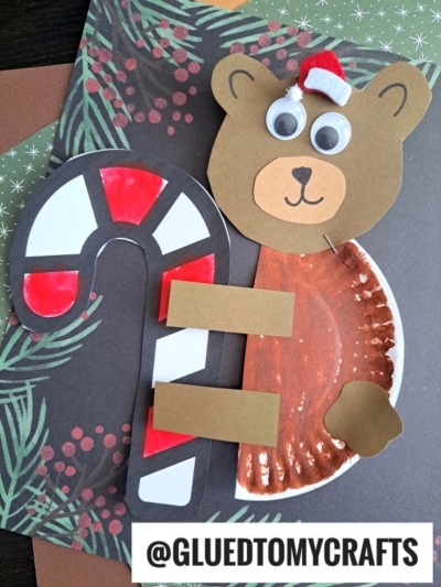 A paper plate Christmas bear with googly eyes and a Santa hat hugs a red and white candy cane, crafted from painted paper and displayed on a festive background.