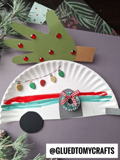 A festive paper craft features a decorated green paper hand as a tree and a half paper plate Christmas camper, painted with colorful lines and lights. A small wreath decorates the camper's door, adding extra holiday cheer.