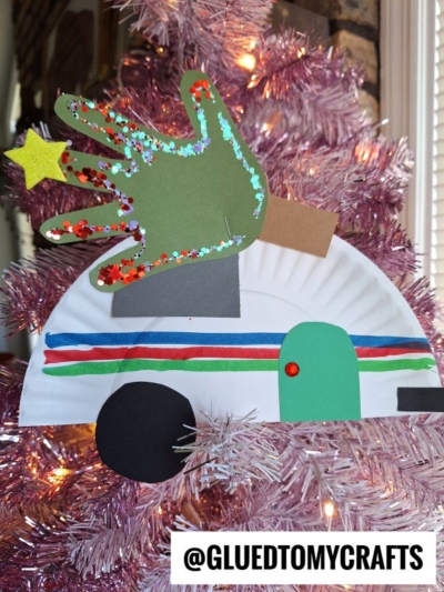 A colorful paper plate Christmas camper craft featuring an RV with a green handprint decorated in glitter, attached to a pink Christmas tree. The RV has multicolored stripes and construction paper details. @GLUEDTOMYCRAFTS is written at the bottom.