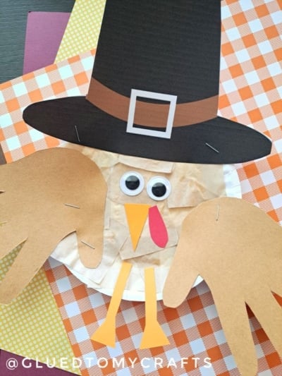 A paper plate pilgrim turkey craft for kids, featuring a construction paper hat, googly eyes, beak, wattle, and handprint wings, displayed on patterned sheets.