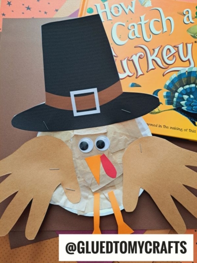 A paper plate pilgrim turkey craft with a black hat, googly eyes, orange beak, and handprint wings is displayed on a brown background next to the book How to Catch a Turkey. @GLUEDTOMYCRAFTS is written at the bottom. Perfect turkey craft for kids!.