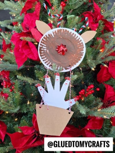 A handmade reindeer ornament hangs on a Christmas tree. The reindeer, inspired by a reindeer hot air balloon, has a brown paper plate face, paper ears, a red bow nose, and handprint cutout body. Red poinsettias brighten the tree.