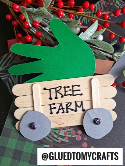 A handmade craft features a popsicle stick Christmas tree wagon with TREE FARM written on it, gray paper wheels, and a green paper handprint tree. Red berries and leaves are in the background. The text @GLUEDTOMYCRAFTS is at the bottom.